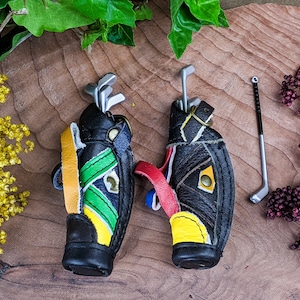 May include: Miniature golf bag keychains in black leather with colorful accents of yellow, green, and red. Each bag holds miniature golf clubs. One golf club is separate. The keychains are on a wooden surface.