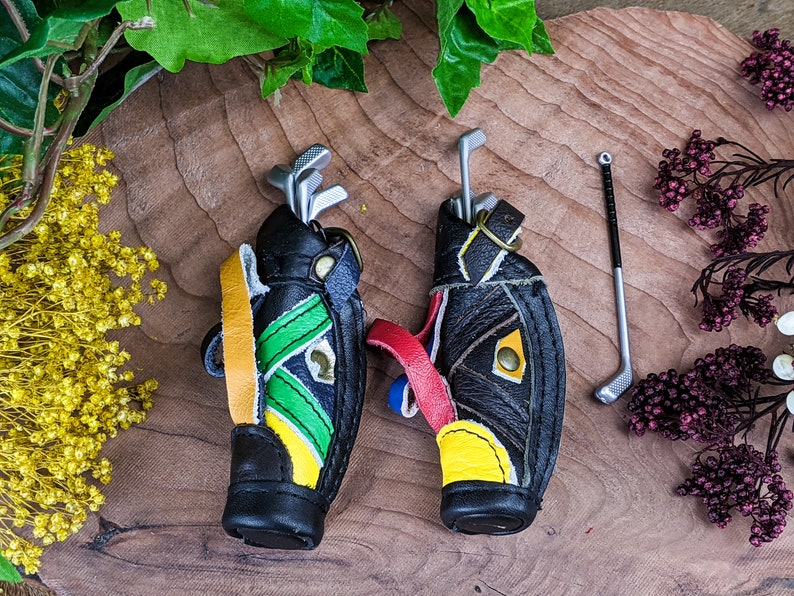 May include: Miniature golf bag keychains in black leather with colorful accents of yellow, green, and red. Each bag holds miniature golf clubs. One golf club is separate. The keychains are on a wooden surface.
