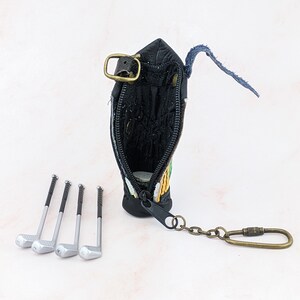 May include: Black leather golf bag-shaped coin purse with a zipper, a metal clasp, and a keychain. Four miniature golf clubs are arranged in front of the purse. The purse has green, yellow, and orange embroidery.