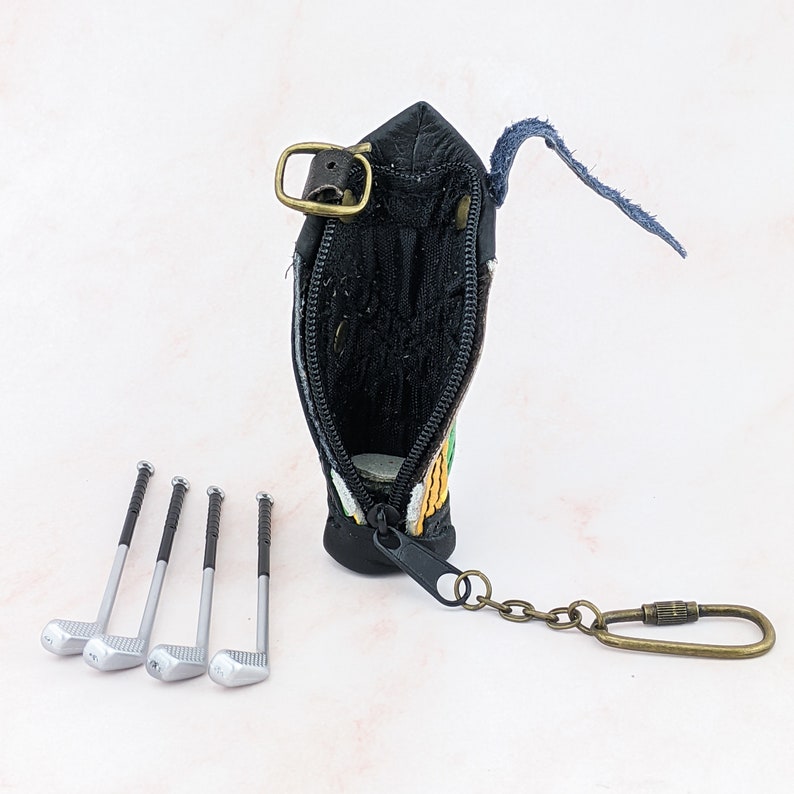 May include: Black leather golf bag-shaped coin purse with a zipper, a metal clasp, and a keychain. Four miniature golf clubs are arranged in front of the purse. The purse has green, yellow, and orange embroidery.