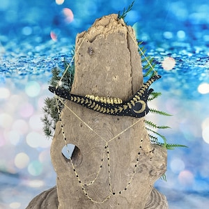 May include: Black and gold painted bone necklace with a crescent moon and stars design. The necklace is hanging on a piece of driftwood with black beads and gold chain.