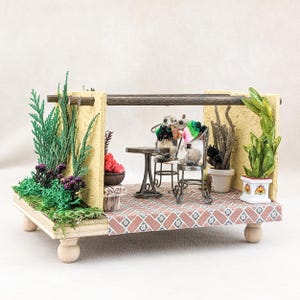 May include: A miniature scene with two small animal figures sitting at a table with drinks, surrounded by potted plants and a wooden structure. The scene is set on a wooden platform with a tiled floor.