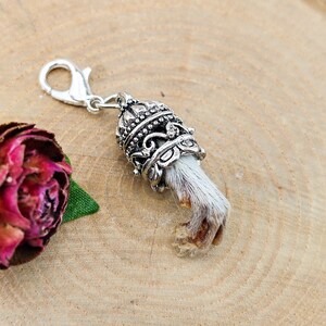 May include: A silver-toned metal charm featuring a lobster claw clasp attached to an ornate, filigree-style cap. The cap holds a small, textured, light-colored animal paw with visible fur and claws, extending downward from the decorative metal setting.