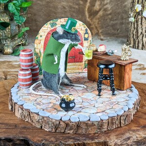 May include: A whimsical diorama featuring a mouse dressed in a green jacket and hat, holding a drink at a miniature bar. The scene includes barrels, a stool, and a pot of gold, all set on a stone-like base, evoking a St. Patrick's Day theme.