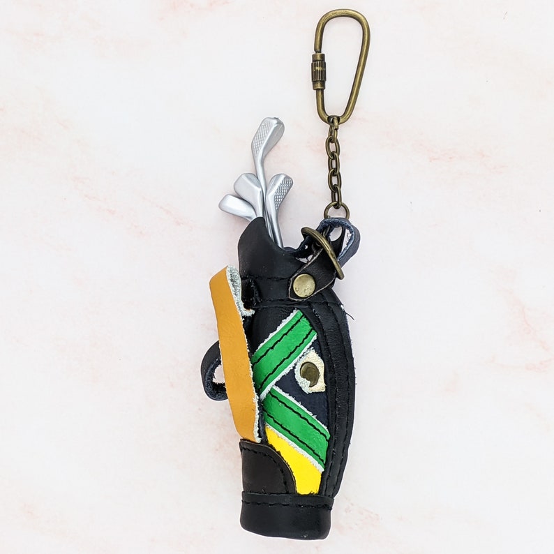 May include: A black leather golf bag keychain with silver golf clubs inside. The bag has green and yellow accents and a tan strap. It hangs from a bronze-colored clasp and chain.