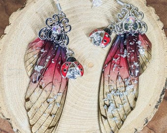 May include: A pair of dangle earrings featuring iridescent, wing-shaped charms with red, purple, and gold hues. Each earring has a silver filigree top with a clear gem and a red ladybug charm with a clear gem.