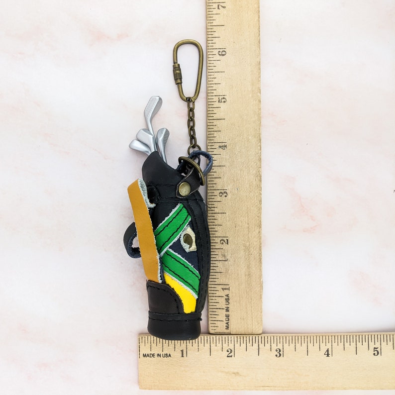 May include: A miniature black golf bag keychain with a gold-colored chain and clasp. The bag features green and yellow accents and contains silver golf clubs. The keychain measures approximately 2 inches in height.