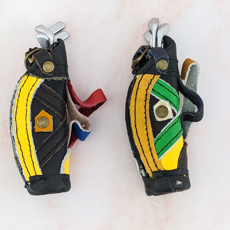 May include: Two miniature golf bag keychains. The bags are black with yellow and green accents, and each has a set of silver golf clubs. The keychains have leather straps in red, blue, and tan.