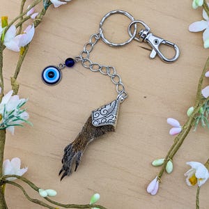 May include: A unique keychain featuring a small animal paw with a decorative silver cap. The keychain includes a silver chain, a blue evil eye charm, and a silver clasp. The paw is brown and has black claws.