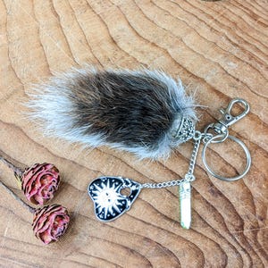 May include: A keychain with a fluffy, gray and brown fur charm, a silver-colored clasp, and a small charm with a sun design. A small, light green crystal is attached to the chain. Two dried, red flowers are in the background.