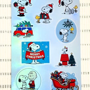 May include: A sheet of Christmas stickers featuring Snoopy and Woodstock in festive scenes. Snoopy is shown wearing a Santa hat, holding gifts, and driving a red sleigh. The stickers include the text "MERRY CHRISTMAS".