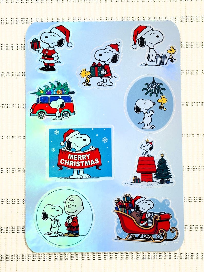 May include: A sheet of Christmas stickers featuring Snoopy and Woodstock in festive scenes. Snoopy is shown wearing a Santa hat, holding gifts, and driving a red sleigh. The stickers include the text "MERRY CHRISTMAS".