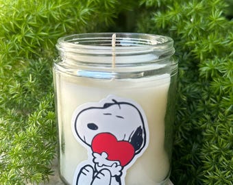 May include: A clear glass jar candle filled with white wax and a single centered wick. A sticker featuring a black and white cartoon dog holding a bright red heart is applied to the front of the glass container, which sits on a light gray surface.