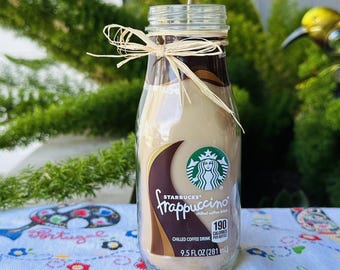 May include: A Starbucks Frappuccino-themed candle in a clear glass bottle. The candle has a light brown color, with a dark brown band at the top and a decorative raffia bow. The label reads "Frappuccino" and "9.5 FL OZ (281 ml)".