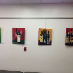 May include: Four framed paintings of wine bottles on a white wall. Each painting features a different arrangement of wine bottles with various labels and colors. The paintings are hung on a metal track system.