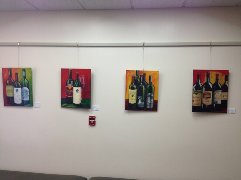 May include: Four framed paintings of wine bottles on a white wall. Each painting features a different arrangement of wine bottles with various labels and colors. The paintings are hung on a metal track system.