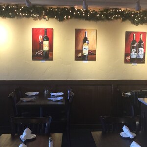 May include: A restaurant interior with four framed paintings of wine bottles on the wall. The paintings are in a modern style and feature a variety of wine bottles. The restaurant is decorated with a rustic theme and has wooden tables and chairs.