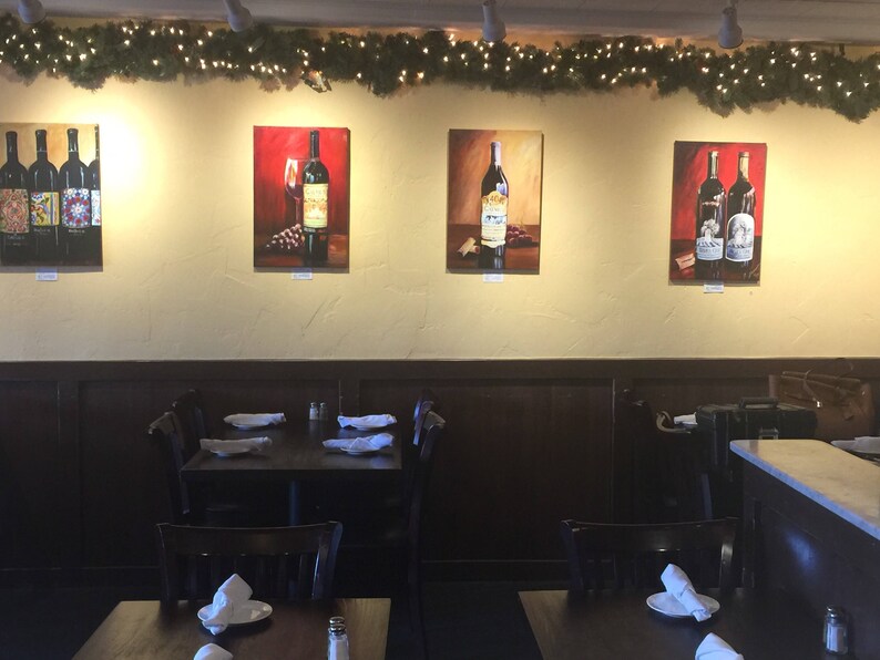 May include: A restaurant interior with four framed paintings of wine bottles on the wall. The paintings are in a modern style and feature a variety of wine bottles. The restaurant is decorated with a rustic theme and has wooden tables and chairs.