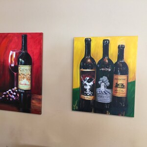 May include: Two framed paintings of wine bottles. The first painting is on a red background with a single bottle of Caymus Vineyards wine and a glass of red wine. The second painting is on a yellow background with three bottles of wine. The bottles are labeled "The Prisoner", "Silver Oak", and "Duckhorn".