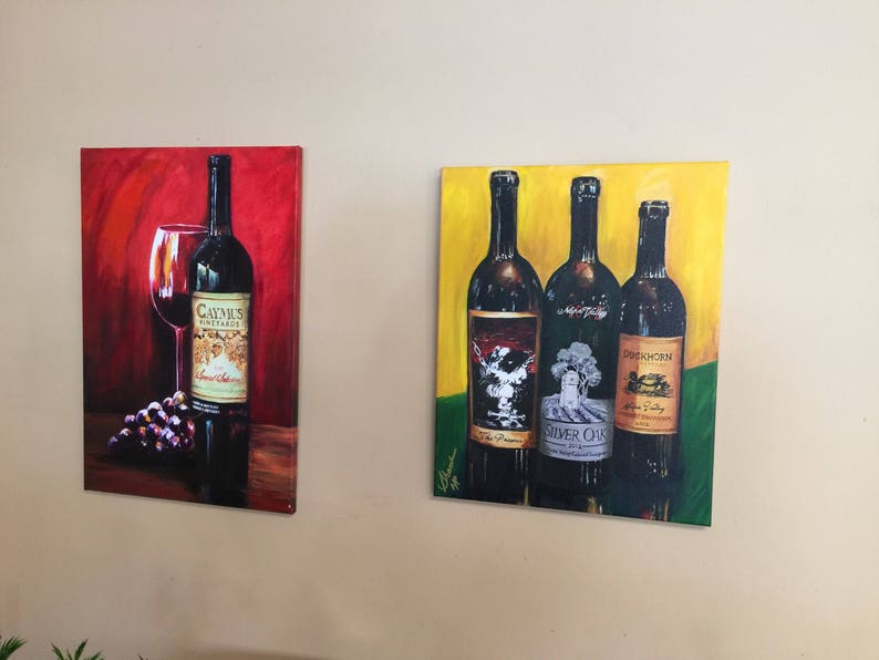 May include: Two framed paintings of wine bottles. The first painting is on a red background with a single bottle of Caymus Vineyards wine and a glass of red wine. The second painting is on a yellow background with three bottles of wine. The bottles are labeled "The Prisoner", "Silver Oak", and "Duckhorn".