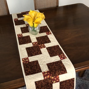 May include: A quilted table runner featuring a repeating pattern of brown squares and cream fabric. The runner is decorated with autumnal fabric squares and is approximately 30 cm wide. A glass vase with yellow flowers sits on top.