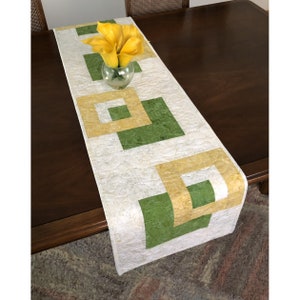 Peut inclure: Un chemin de table blanc avec un motif géométrique de carrés verts et jaunes. Le chemin de table est sur une table en bois avec un vase de fleurs jaunes.