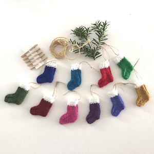 May include: A selection of small, knitted Christmas stockings in diverse colours, such as blue, green, red, and purple, each with a white cuff. They are displayed with twine, wooden pegs, and evergreen sprigs, hinting at festive decorations.