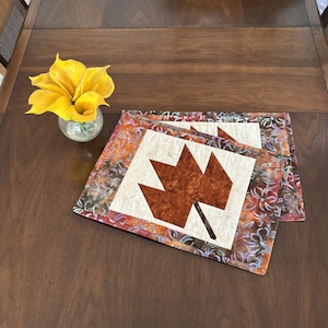 Peut inclure: Deux sets de table matelassés avec un motif de feuille d'érable brun sur fond blanc. Les sets de table ont une bordure à motif floral brun, violet et blanc.