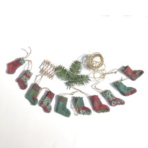 May include: A garland of ten knitted red and green Christmas stockings with white accents. The stockings are strung together with twine and are hanging on a white background.