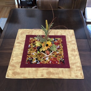 Peut inclure: Un chemin de table carré avec une bordure bordeaux et or et un panneau central imprimé de feuilles d'automne. Le chemin de table est décoré d'un petit arrangement de feuillage d'automne et de baies.