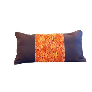 Puede incluir: Un cojín decorativo rectangular con un cuerpo marrón oscuro y una banda central de tela naranja. La banda naranja presenta un estampado floral en tonos amarillos, naranjas y rojos. El cojín está diseñado para la decoración del hogar.