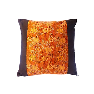May include: Square decorative pillow with a floral pattern in shades of orange, yellow, and brown. The center panel features a detailed flower design, framed by solid dark brown fabric on the sides. The pillow is square and appears to be made of fabric.