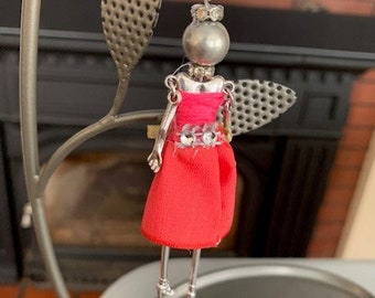 May include: A silver pendant necklace featuring a small doll figure wearing a red dress with silver embellishments. The doll has a silver head with rhinestones for eyes.