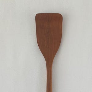 May include: A wooden spatula with a rectangular head and a long, slender handle. The spatula is made of a light brown wood and is designed for cooking and food preparation. The spatula is shown against a white background.
