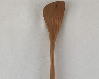 May include: A wooden spatula with a curved, asymmetrical head and a long, slender handle. The spatula is made of light brown wood, showcasing the natural grain. The kitchen utensil is designed for cooking and food preparation.