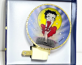May include: A Betty Boop night light in a blue box. The night light features a circular design with Betty Boop in a red dress, standing in front of a yellow moon and stars. The night light has a gold-colored trim and a plug.