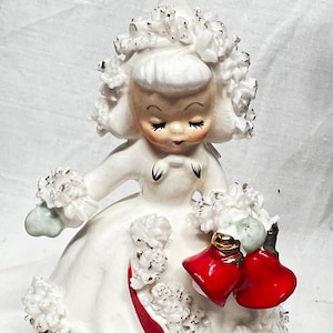 May include: A vintage ceramic figurine of a girl in a white dress with a red ribbon. The figurine has a white floral headpiece and holds two red heart-shaped ornaments. The girl's face is painted with closed eyes and rosy cheeks.