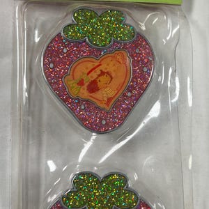 May include: Two glittery strawberry-shaped stickers featuring Strawberry Shortcake. Each sticker has a pink glitter base, a green glitter top, and an orange heart-shaped center with a Strawberry Shortcake illustration.