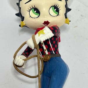 May include: A figurine of a cartoon character wearing a white cowboy hat, red and black plaid shirt, blue jeans, and a brown rope. The character has green eyes, red lips, and black hair.
