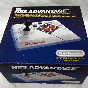 May include: A blue box featuring the Nintendo NES Advantage controller. The controller is gray with a black joystick and red buttons. The box has the text "NES ADVANTAGE" and "Nintendo ENTERTAINMENT SYSTEM".