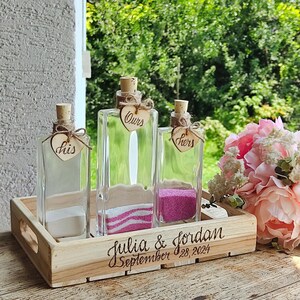 Peut inclure: Trois bouteilles en verre avec des bouchons en liège et des étiquettes en forme de cœur en bois qui disent "his", "hers" et "ours". Les bouteilles sont remplies de sable et reposent sur un plateau en bois avec le texte "julia & jordan September 28, 2024" gravé dessus.