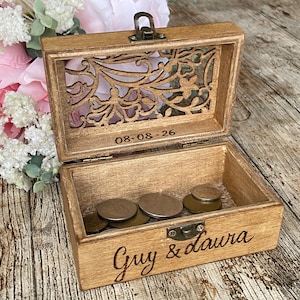 May include: A rectangular wooden box with a hinged lid featuring a laser-cut floral pattern. The interior lid is engraved with the date 08-08-26. The front of the box is engraved with the text Guy & Laura. Several metal coins rest inside.