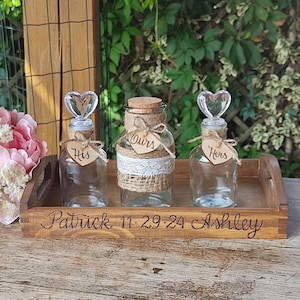 May include: Three glass jars with cork tops and heart-shaped stoppers on a wooden tray. The jars are decorated with burlap and lace. The jars are labeled "His", "Ours", and "Hers". The tray is engraved with the names "Patrick 11.29.24 Ashley".