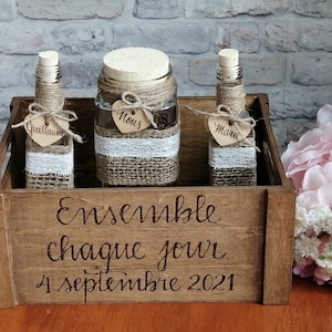 May include: A wooden crate holding three glass bottles and a jar, decorated for a wedding. The bottles and jar are wrapped with twine, burlap, and lace. The crate has the words "Ensemble chaque jour 4 septembre 2021" engraved on it.