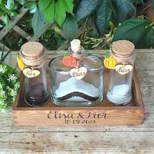 Peut inclure: Une boîte en bois contenant trois bocaux en verre pour une cérémonie de sable. Les bocaux sont étiquetés "Lui", "Nous" et "Elle" avec de petites citrouilles en bois et des cœurs. La boîte est gravée avec "Elisa & Pier 10.09.2023".