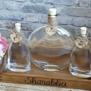 Könnte beinhalten: Drei klare Glasflaschen mit Korkstopfen und Kordel-Akzenten auf einem Holztablett. Jede Flasche hat ein herzförmiges Etikett mit einem Buchstaben und einem Datum. Das Tablett ist mit dem Wort "Sharabkha" graviert.