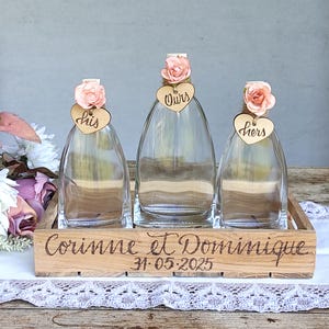 May include: Three clear glass bottles with cork stoppers, each adorned with a peach-colored fabric flower and a small wooden heart tag labeled his, ours, and hers. The bottles sit in a wooden crate engraved with Corinne et Dominique 31.05.2025.