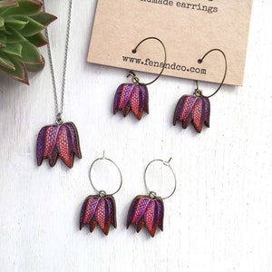 May include: A set of handmade earrings and a necklace featuring a purple and red flower design. The earrings are hoop style and the necklace has a silver chain. The jewelry is displayed on a white surface with a brown card that reads "Fen & Co Handmade earrings www.fenandco.com".