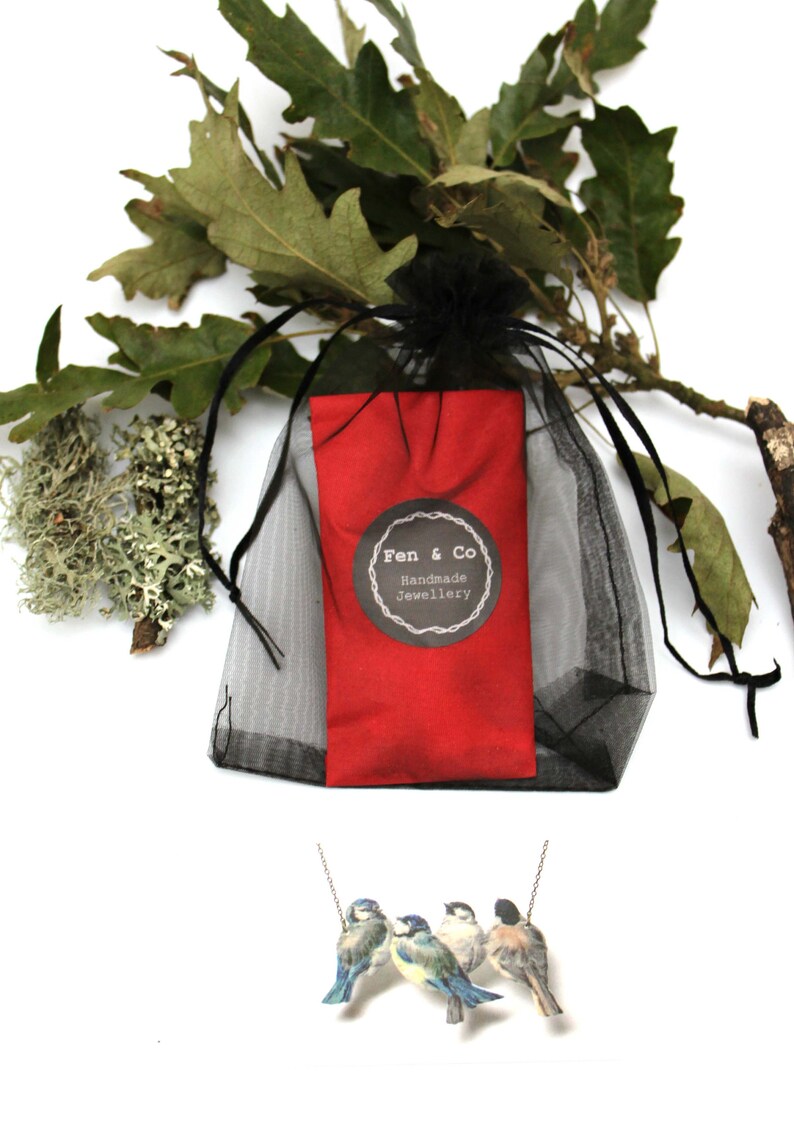 May include: A silver necklace with four bluebird charms. The necklace is packaged in a red pouch with a black mesh bag. The pouch has a black and white label that reads "Fen & Co Handmade Jewellery".