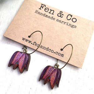 May include: A pair of handmade earrings featuring a purple and orange flower design. The earrings are made of wood and have a bronze hoop.